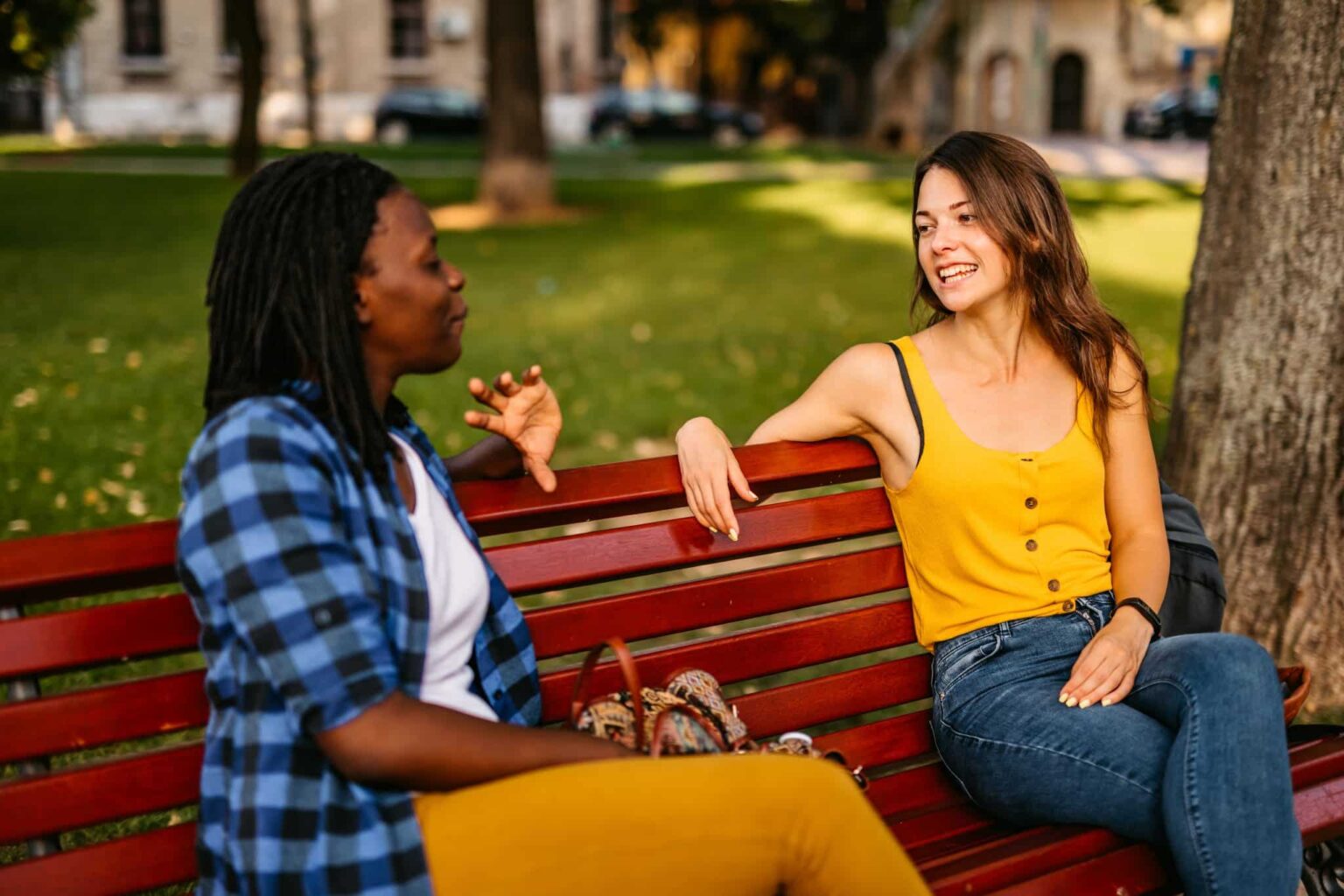 after aphasia treatment Two woman sit on a park bench having a discussion. The woman on the left is mid sentence and the woman on the right is watching her and smiling.